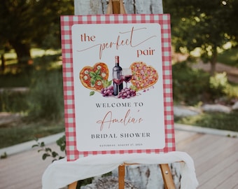 May include: A white sign with red and white checkered border. The sign says "the perfect pair" with a watercolor illustration of a heart-shaped pizza, a bottle of wine, a glass of wine, and grapes. The sign also says "Welcome to Amelia's Bridal Shower" and "Saturday, August 27, 2022".