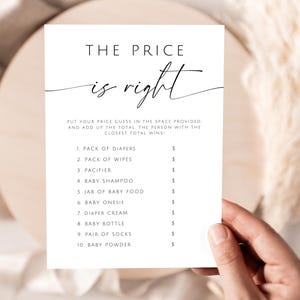 May include: A white card with the text "THE PRICE is right" in elegant script. Below, a list of baby items with spaces for price guesses. The card is held by a hand, suggesting a baby shower game.