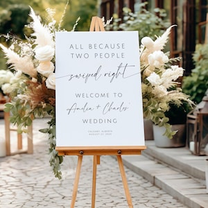 May include: A white wedding sign on a wooden easel, with the text "ALL BECAUSE TWO PEOPLE swiped right WELCOME TO Amelia + Charles WEDDING" in black script. The sign is surrounded by floral arrangements with white roses and pampas grass.