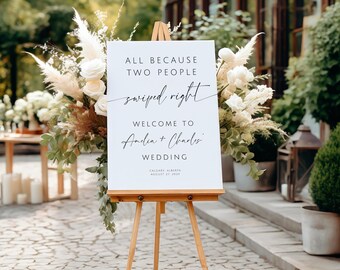 Puede incluir: Un letrero de boda blanco sobre un caballete de madera, con el texto "ALL BECAUSE TWO PEOPLE swiped right WELCOME TO Amelia + Charles WEDDING" en escritura negra. El letrero está rodeado de arreglos florales con rosas blancas y hierba de la Pampa.