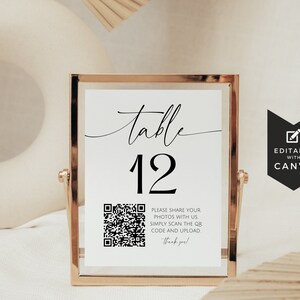 May include: A white table number sign with black text and a QR code. The sign reads "table 12" and includes instructions to share photos by scanning the QR code. The sign is in a gold frame and is sitting on a white surface.