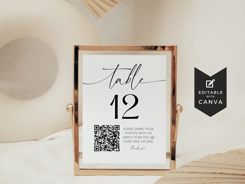 May include: A white table number sign with black text and a QR code. The sign reads "table 12" and includes instructions to share photos by scanning the QR code. The sign is in a gold frame and is sitting on a white surface.