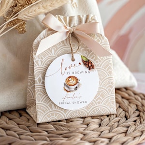 May include: A beige gift bag with a cream-colored ribbon and a round tag. The tag says "Love is Brewing" with a coffee cup illustration and "Amelia's Bridal Shower" text. The bag is on a woven mat.