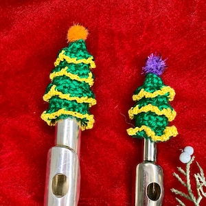 Puede incluir: Dos flautas de plata decoradas con árboles de Navidad de crochet verde y amarillo. El árbol más grande tiene un pompón naranja en la parte superior y el árbol más pequeño tiene un pompón morado en la parte superior.
