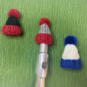 Puede incluir: Tres gorros de punto de diferentes colores y patrones. Los gorros son rojo y negro, rojo y gris, y blanco y azul. Los gorros están sobre una superficie verde.