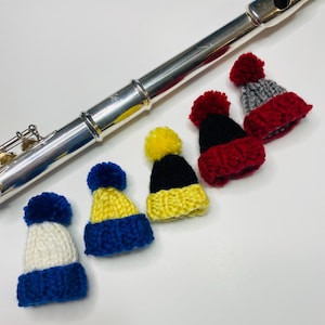Puede incluir: Cinco gorros de punto en miniatura en varios colores, incluyendo rojo, azul, amarillo, blanco y gris, con pompones en la parte superior. Los gorros están dispuestos sobre una superficie blanca junto a una flauta de plata.