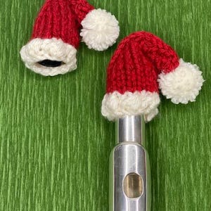 May include: Two red and white knitted Santa hats with white pom-poms are placed on top of a silver flute.