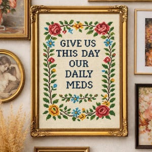 May include: A cross-stitch embroidery in a gold frame featuring the text GIVE US THIS DAY OUR DAILY MEDS in dark blue letters, surrounded by a rectangular border of green vines, red roses, and small yellow and blue flowers on cream fabric.