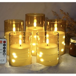 May include: A set of five cylindrical LED candles encased in amber-tinted glass sleeves, each featuring a warm white flame and internal fairy lights. A white remote control with various timer and light mode buttons is positioned to the left.