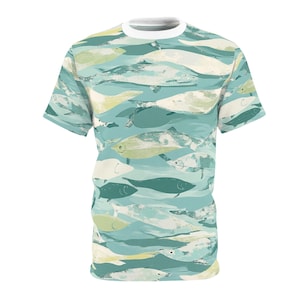 May include: A short-sleeved t-shirt with a white collar. The shirt features a repeating pattern of fish in shades of blue, green, and cream. The fish are stylized with brushstroke-like details, creating a textured appearance. The overall design has a nautical theme.
