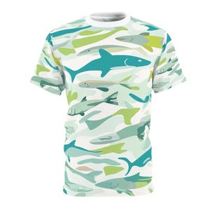 May include: White crew neck t-shirt with a repeating pattern of various fish in shades of teal, green, and gray. The fish are stylized and arranged on a white background, creating a camouflage effect. The shirt has short sleeves.
