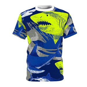 May include: A short-sleeved t-shirt featuring a vibrant shark pattern. The design includes neon yellow, blue, gray, and white shark illustrations on a dark blue background. The shirt has a white crew neck and blue sleeves.
