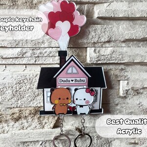 Puede incluir: Un juego de llaveros decorativos de acr&iacute;lico con un dise&ntilde;o de casa, dos personajes de dibujos animados y un grupo de globos en forma de coraz&oacute;n. El juego incluye dos llaveros y un llavero. El texto "Dudu Bubu" es visible.