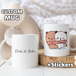 May include: Two white ceramic mugs. One mug has the text "Dudu & Bubu" in black script. The other mug features a cartoon illustration of two bears wearing a scarf. The image also includes the text "+ Stickers".