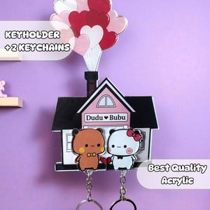 May include: A keyholder with two keychains, shaped like a house with a chimney and heart-shaped balloons. Two cartoon bear characters are featured below the house. The text "Dudu Bubu" is visible.