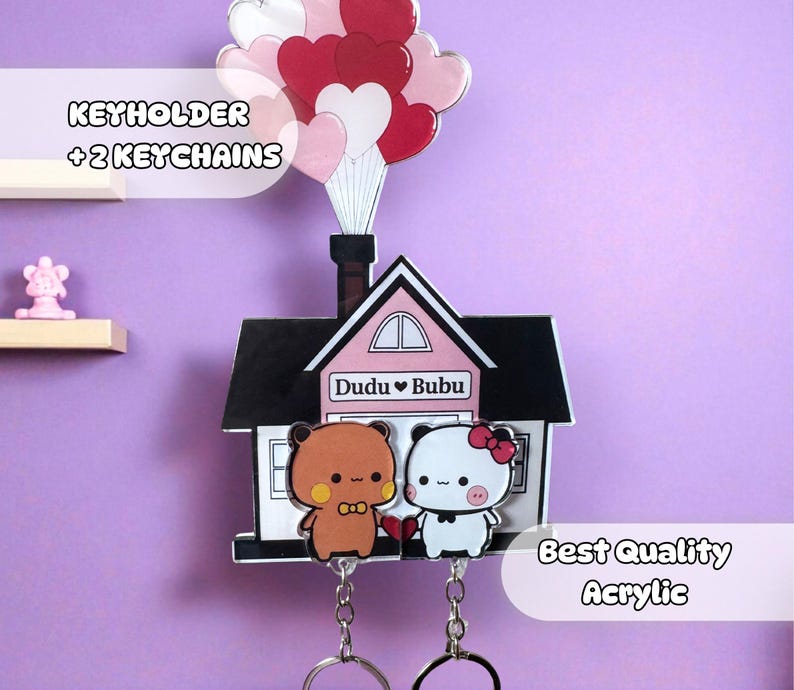Puede incluir: Un llavero decorativo con dos llaveros. El llavero tiene forma de casa con chimenea y globos en forma de coraz&oacute;n. Dos personajes de osos de dibujos animados aparecen debajo.