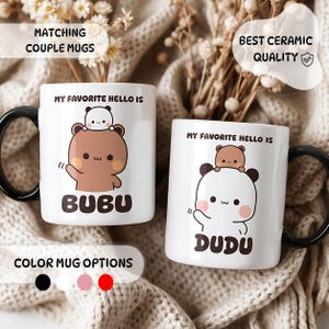 Op de afbeelding: Twee witte keramische mokken met zwarte oren en cartoon beer illustraties. De linker mok toont een bruine beer met de tekst MY FAVORITE HELLO IS BUBU. De rechter mok toont een witte beer met de tekst MY FAVORITE HELLO IS DUDU.