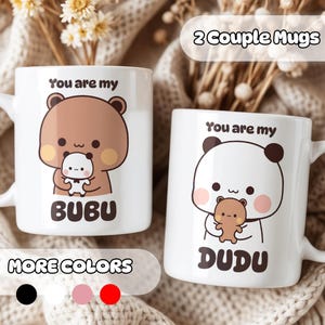 May include: Two white ceramic mugs featuring cartoon bear illustrations. The left mug shows a brown bear holding a small white bear with the text "You are my BUBU". The right mug shows a white bear holding a small brown bear with the text "You are my DUDU".