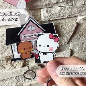 Puede incluir: Un llavero con dos personajes de dibujos animados, un oso pardo y un panda blanco con un lazo rosa. El llavero est&aacute; unido a un charm con forma de casa con el texto "Dudu Bu". El tama&ntilde;o es de unos 10-14 cm.