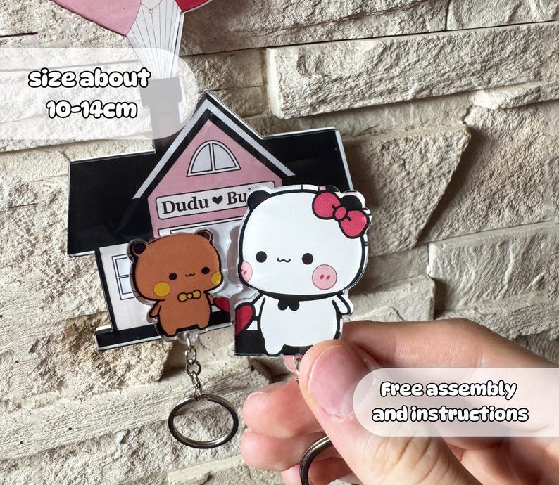 Puede incluir: Un llavero con dos personajes de dibujos animados, un oso pardo y un panda blanco con un lazo rosa. El llavero est&aacute; unido a un charm con forma de casa con el texto "Dudu Bu". El tama&ntilde;o es de unos 10-14 cm.
