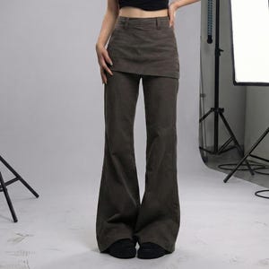 May include: A person wearing a black short-sleeved crop top and olive green flared pants. The pants have a unique skirt-like overlay at the waist. The outfit is styled with black shoes. The background is a studio setting with lighting equipment.
