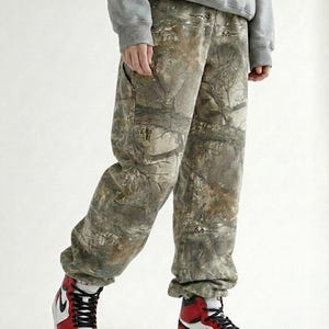 May include: A person wearing a gray hoodie, camouflage pants, and red, white, and black high-top sneakers. The pants have a woodland pattern. The sneakers have a white base with red and black accents.