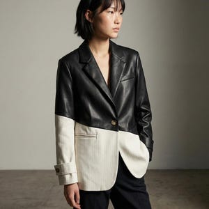 Peut inclure: Un blazer bicolore, avec une partie supérieure en cuir noir et une section inférieure crème à rayures verticales. Le blazer a une fermeture à un seul bouton et un revers cranté. Les manches sont retroussées et le vêtement est associé à un pantalon noir.