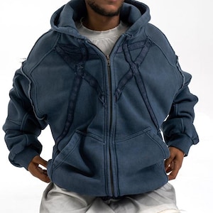 May include: A blue zip-up hoodie with a distressed design. The hoodie has a hood, pockets, and a zip. The model is wearing light-wash ripped jeans and futuristic sunglasses. The background is plain white.
