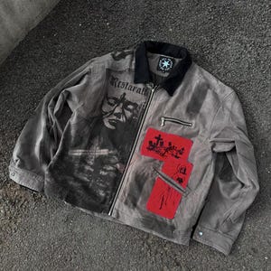 Könnte beinhalten: Graue Jacke mit schwarzem Kragen und Reißverschluss. Die Jacke hat einen grafischen Aufdruck auf der Vorderseite und zwei rote Aufnäher mit Illustrationen. Der Text "Restarall" ist sichtbar.
