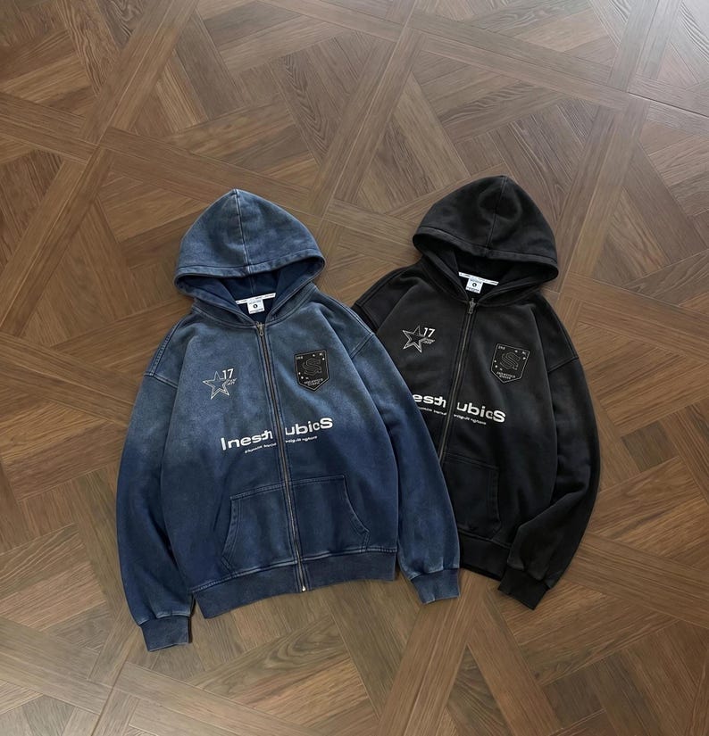 May include: Two zip-up hoodies, one in faded blue and the other in black, with a star and shield emblem. The blue hoodie has the text "Inest UbicS" printed on the front. Both hoodies have a hood and front pockets.