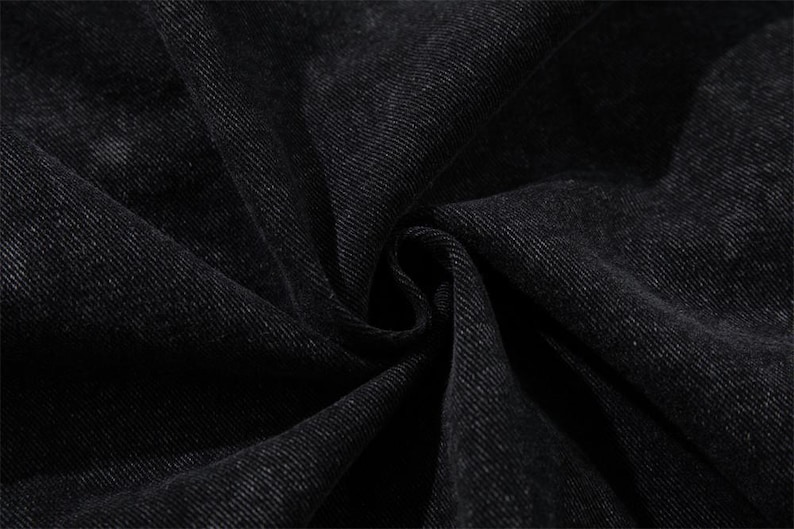 May include: Close-up of black denim fabric with visible texture and folds. The dark material has a subtle sheen, suggesting a garment or textile product. The image highlights the weave and color variations of the fabric.