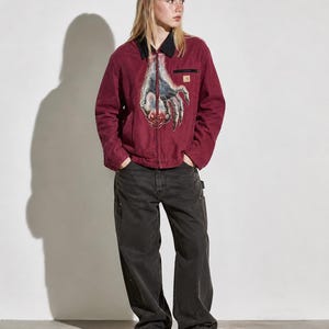 May include: A burgundy work jacket with a black collar and a graphic of a hand on the front. The jacket has a small pocket on the chest and is paired with black work pants and black shoes.