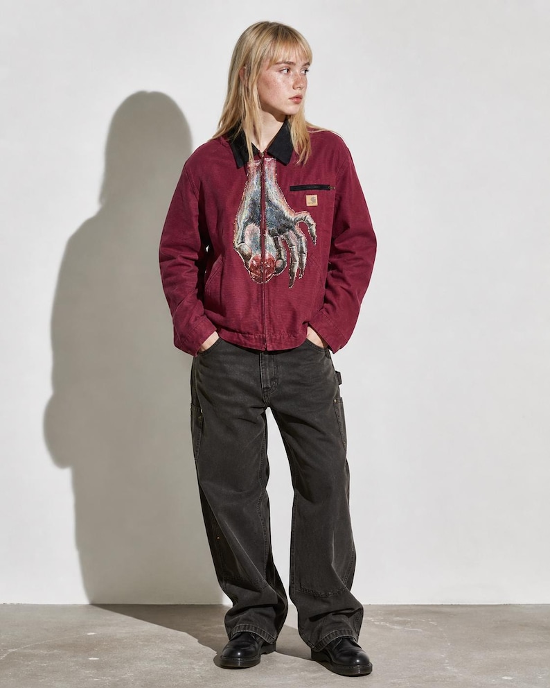 May include: A burgundy work jacket with a black collar and a graphic of a hand on the front. The jacket has a small pocket on the chest and is paired with black work pants and black shoes.