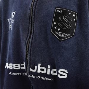 May include: A navy blue zip-up hoodie with white embroidered text and patches. The hoodie features a star graphic and the text "17" on the left chest. The right chest has a black patch with the text "INS" and "INSSTUDIOS FIERCE".