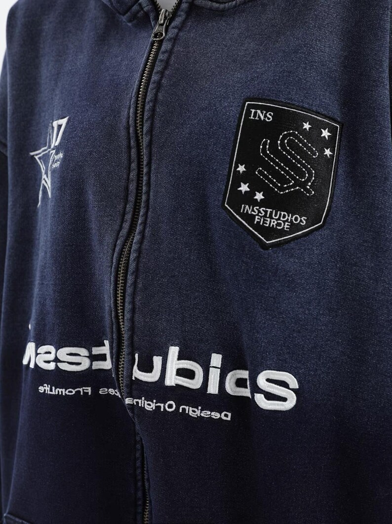 May include: A navy blue zip-up hoodie with white embroidered text and patches. The hoodie features a star graphic and the text "17" on the left chest. The right chest has a black patch with the text "INS" and "INSSTUDIOS FIERCE".