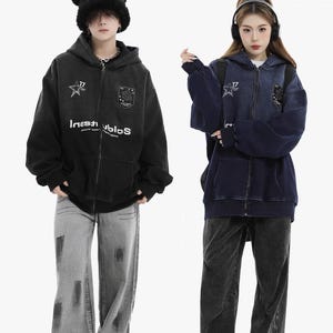 May include: Two people wearing matching zip-up hoodies and jeans. One hoodie is black, the other is navy blue. Both hoodies have a star and shield emblem, and the text "Investiblos". The jeans are gray and black.