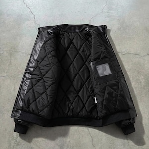 May include: Black leather jacket with a quilted interior. The jacket is open, revealing the diamond-patterned lining and a small pocket. The cuffs and hem are ribbed. The jacket is displayed against a gray concrete background.