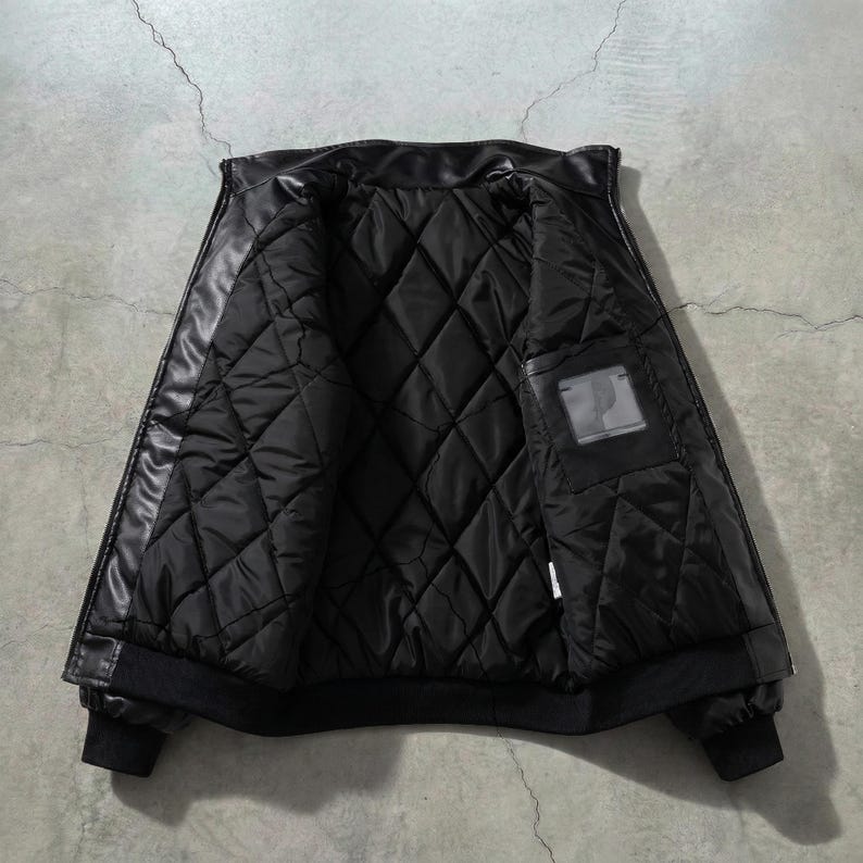 May include: Black leather jacket with a quilted interior. The jacket is open, revealing the diamond-patterned lining and a small pocket. The cuffs and hem are ribbed. The jacket is displayed against a gray concrete background.