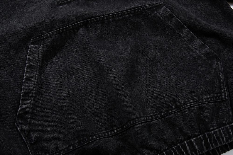 May include: Close-up of a black denim hoodie pocket. The pocket is large and rectangular, with visible stitching around the edges. The fabric has a slightly faded appearance, suggesting a worn or washed look. The hoodie's elastic waistband is visible.