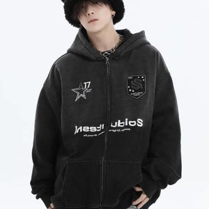 May include: Black zip-up hoodie with a faded wash effect. Features a star graphic with the number 17, and a shield emblem. The hoodie has the word "INSTUDIOS" printed in white, and is paired with a black bear hat.