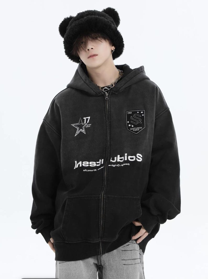 May include: Black zip-up hoodie with a faded wash effect. Features a star graphic with the number 17, and a shield emblem. The hoodie has the word "INSTUDIOS" printed in white, and is paired with a black bear hat.