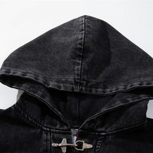 May include: Close-up of a black denim hooded jacket. The jacket features a hood, a metal toggle closure, and a faded, distressed appearance. The fabric appears to be a dark wash denim.