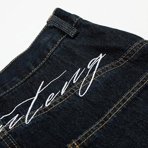 K&ouml;nnte beinhalten: Nahaufnahme einer dunkelblauen Jeans mit wei&szlig; gesticktem Text. Die Jeans hat sichtbare gelbe N&auml;hte und ein klassisches Design. Der Text scheint ein Markenname oder ein Designelement zu sein.