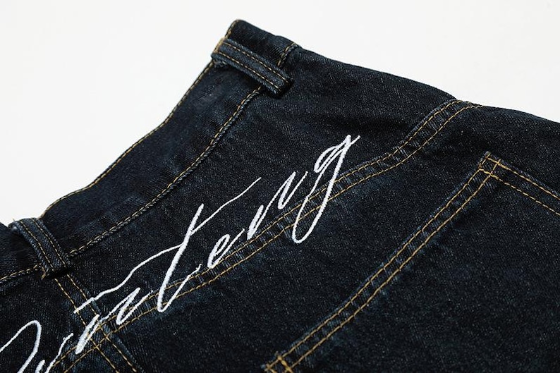 K&ouml;nnte beinhalten: Nahaufnahme einer dunkelblauen Jeans mit wei&szlig; gesticktem Text. Die Jeans hat sichtbare gelbe N&auml;hte und ein klassisches Design. Der Text scheint ein Markenname oder ein Designelement zu sein.