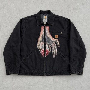 May include: Black jacket with a full-zip closure, collared neckline, and two front pockets. The jacket features a graphic of a hand with a red and gray design on the front. A small logo patch is on the chest.