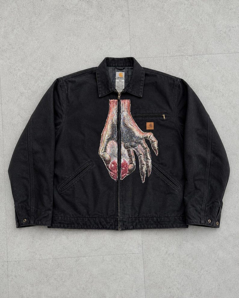 May include: Black jacket with a full-zip closure, collared neckline, and two front pockets. The jacket features a graphic of a hand with a red and gray design on the front. A small logo patch is on the chest.