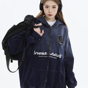 May include: A navy blue zip-up hoodie with a faded denim effect. The hoodie features a white graphic print that reads "Inest Ubios" and a patch on the upper chest. The model is wearing headphones and carrying a black backpack.