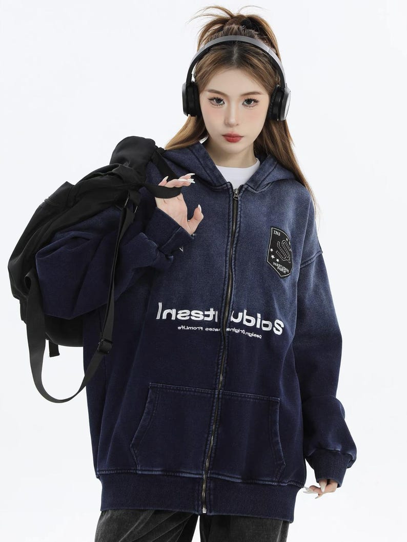 May include: A navy blue zip-up hoodie with a faded denim effect. The hoodie features a white graphic print that reads "Inest Ubios" and a patch on the upper chest. The model is wearing headphones and carrying a black backpack.