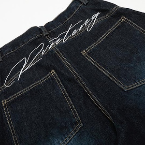 K&ouml;nnte beinhalten: Dunkelblaue Jeans mit wei&szlig;em, gesticktem Text am hinteren Bund. Die Jeans hat zwei Ges&auml;&szlig;taschen mit gelben N&auml;hten. Der Denim hat unten eine leichte Ausbleichung.