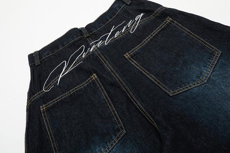 K&ouml;nnte beinhalten: Dunkelblaue Jeans mit wei&szlig;em, gesticktem Text am hinteren Bund. Die Jeans hat zwei Ges&auml;&szlig;taschen mit gelben N&auml;hten. Der Denim hat unten eine leichte Ausbleichung.